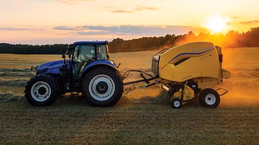 New Holland tractor pulling a Roll-Belt round baler at sunset